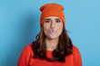 © sementsova321 - Young lady blowing bubble gum isolated over blue background, happy girl with dark hair with burst bubble of gum in mouth, wearing orange jumper and cap, looking at camera, looks funny.