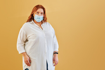 serious redhead female in casual wear in medical protective mask looking at camera alone isolated over yellow background. coronavirus, covid-19 concept