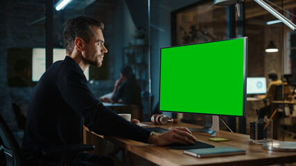 Wall Mural - Young Handsome Specialist Working on Desktop Computer with Green Screen Mock Up Display in a Busy Creative Office with Colleagues. Male Manager with Trimmed Beard is Wearing a Casual Black Jumper.