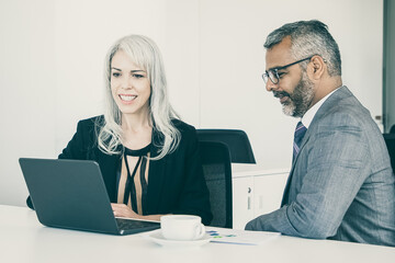 Wall Mural - Two happy colleagues using laptop for video call, sitting at table with cup of coffee, looking at display and talking. Online communication concept