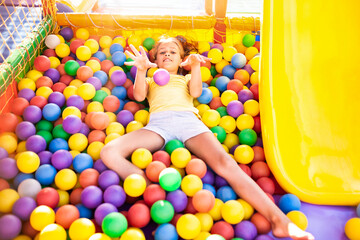 A cute cheerful girl lies on the playground with soft and bright equipment and throws colorful balloons into the camera, enjoying the warm summer sun