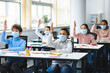 © Prostock-studio - Schoolchildren raising hands at classroom, wearing medical masks