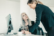 © Mangostar - Project manager affixing signature on employees report. Female business colleagues sitting and standing at workplace with monitor and coffee cup. Business communication concept