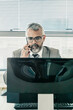 © Mangostar - Serious grey haired businessman making call on cell phone while using computer at workplace in office. Front view. Communication and multitasking concept