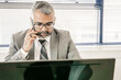 © Mangostar - Focused mature executive speaking on mobile phone while using computer at workplace in office. Front view. Digital communication and multitasking concept