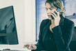 © Mangostar - Serious female professional talking on mobile phone while using computer at workplace in office. Medium shot. Digital communication and multitasking concept