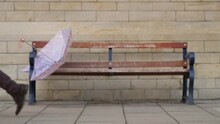 Defocused Medium Shot Of Discarded Umbrella On Bench As People Walk Past