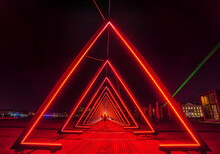 Series Of Triangular Gates Of Light Or Intense Red Light Tunnel. The Gate Of Light Installation Consists Of A Succession Of Gates (triangles) Where Blurred People Look Across An Extraterrestrial World