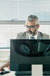 © Mangostar - Serious grey haired businessman sitting at workplace with pc monitor and taking cellphone from desk. Front view. Communication and multitasking concept