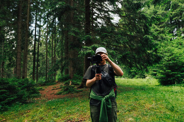 Wall Mural - A male videographer with a camera on a stabilizer stands in the woods against a background of trees and adjusts the technique for shooting. Creating video content in nature