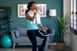 Sporty african young woman exercising on smart stationary bike while drinking water at home.