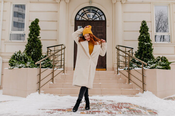 Wall Mural - Blissful ginger woman in white coat posing near home. Outdoor shot of beautiful shy lady enjoying winter.