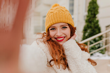 Wall Mural - Outdoor photo of romantic ginger woman wears yellow knitted hat. Good-humoured caucasian girl making selfie in winter.