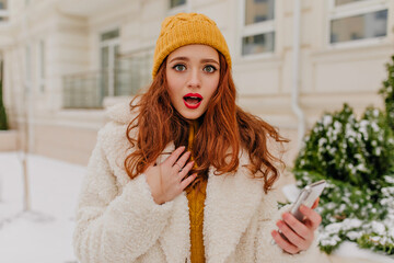 Wall Mural - Surprised ginger girl standing on the street in cold day. Magnificent young woman with phone posing outdoor.