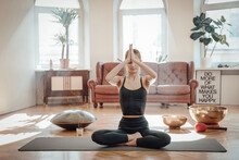 Peaceful And Warm Living Room With Styled Furniture And Caucasian Woman Relaxes Doing Yoga Sitting On Carpet On Floor In Daytime.