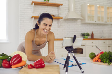 Wall Mural - Fitness blogger, online nutritiologist, diet concept. Smiling slim fitness woman in sportswear standing in kitchen with healthy raw vegetable ingredients and recording video content for blog or vlog