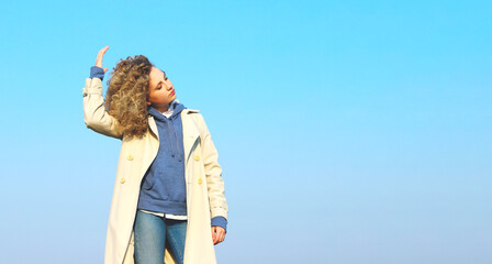 Curly young girl wearing a beige coat, blue jeans and hoodie standing over blue sky background. People, landscapes, travel concept.