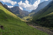 Hikers In Lush Valley Below The Towering Escarment Of The Drakensberg Mountains.
