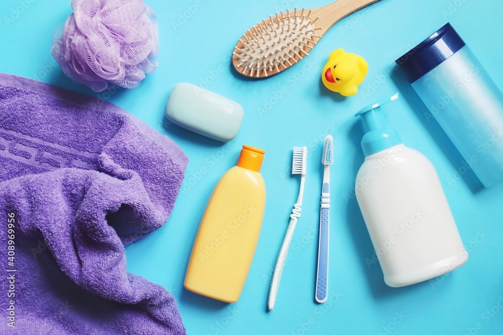 Flat lay composition bath products on a blue background. Purple towel, shampoo bottle, sponge, soap bar, shower gel, hair balm, comb, toothbrushes and rubber duck. Toiletries set - obrazy, fototapety, plakaty 