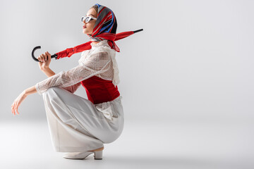 Wall Mural - full length of young woman in headscarf and sunglasses holding red umbrella while sitting on white