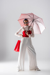 Poster - full length of stylish woman in headscarf and sunglasses holding red umbrella and shopping bag while posing on white