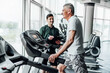 Portrait of an older man on the treadmill, with his coach standing next to him and watching and recording the results. Rehabilitation. Training plan, Coach and patient