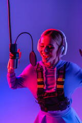 Poster - happy woman in wireless headphones recording song while singing in microphone on purple with color filter