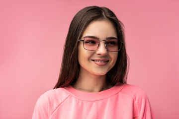 Photo of attractive young woman in glasses, cute face and smiles. Wears casual pink t-shirt white pants isolated pink color background