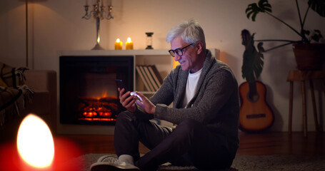 Happy senior man using smartphone while relaxing at home near fireplace