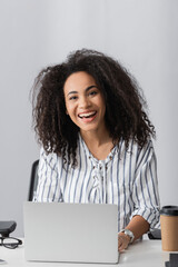 Wall Mural - happy african american freelancer smiling while looking at camera near laptop on desk