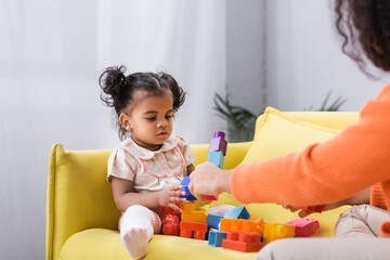 Wall Mural - african american toddler girl sitting on sofa and playing building blocks with mother on blurred foreground