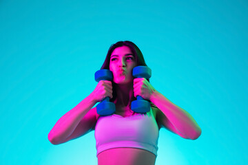 Wall Mural - Lovely. Brunette woman's portrait on blue studio background in mixed neon. Beautiful model training with dumbbells inspired. Concept of human emotions, facial expression, sales, ad, fashion, sport.