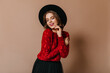 © Look! - Charming caucasian woman in black hat smiling with closed eyes. Studio shot of carefree young lady isolated on brown background.