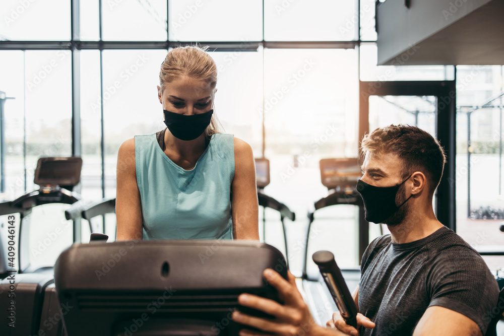 Young fit and attractive woman at body workout in modern gym together with her personal fitness instructor or coach. They keeping distance and wearing protective face masks. Coronavirus sport theme. - obrazy, fototapety, plakaty 