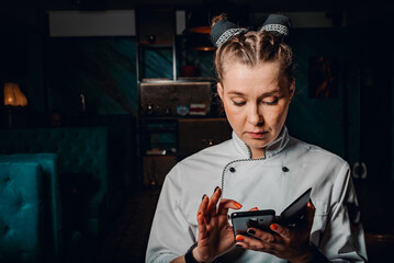 young woman chef with phone in hands on dark background in cafe