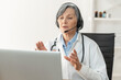 © Vadim Pastuh - Portrait of a senior doctor with a stethoscope in a lab coat sitting at the desk, wearing a headset and looking at the laptop, giving consultation to a client online, working remotely from home