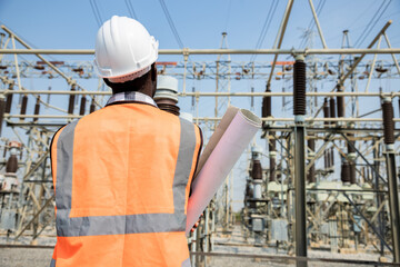 View form back of handsome engineering man holding paper projects plan and wear hardhat in front of High power power plant. Back view of contractor on background of power plant buildings.