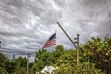 Wall Mural - Building crane used to fly giant American flag about roofs and treetops with downtown skyscraper in distance - cloudy dramatic sky and selective focus