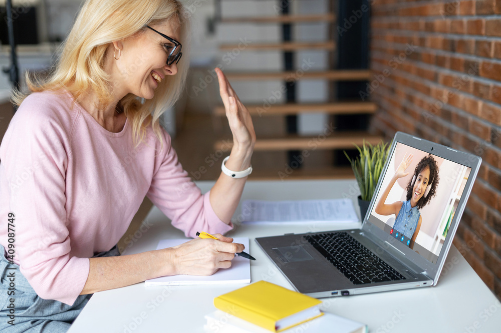 Online teacher, mentor conducts webinar, video lesson. Middle-aged female tutor greeting with a school-age African girl on the laptop screen. E-study concept - obrazy, fototapety, plakaty 