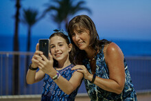 Portrait Of Mother And Teenage Daughter Wearing Blue Dresses And Taking A Selfie With Their Smartphone At Night With A Beach In The Background. Vacation Concept
