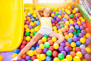 a funny girl sits in a playground with soft and bright equipment and throws colorful balls towards t