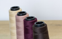 Four Sewing Threads With Different Shades On Their Spools Lined Up Diagonally On A Table With A White Background. The Lines Of Black, Purple, Pink And Yellow Colors. Used In A Sewing Machine.