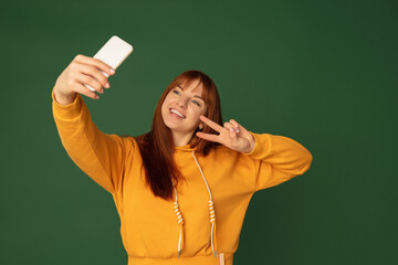 Wall Mural - Selfie, smiling. Caucasian woman's portrait isolated on green studio background with copyspace. Beautiful female model with phone. Concept of human emotions, facial expression, sales, ad, fashion.