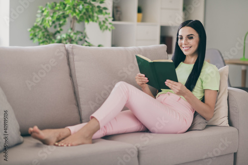 Photo of cheerful clam young lady lay sofa pillows hold book read barefoot wear glasses green t-shirt pink trousers indoors