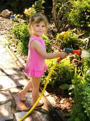 Wall Mural - Beautiful little toddler girl watering garden flowers with water hose on summer day. Happy child helping in family garden, outdoors, having fun with splashing