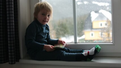 Canvas Print - Toddler child, sitting on the window, watching the snow falling, reading little book