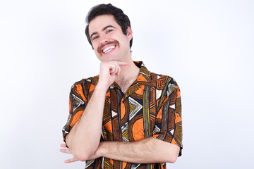 Wall Mural - Optimistic Young caucasian man wearing generic pattern printed shirt against white wall keeps hands partly crossed and hand under chin, looks at camera with pleasure. Happy emotions concept.