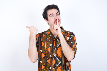 Young caucasian man wearing generic pattern printed shirt against yellow wall asking to be quiet with finger on lips pointing with hand to the side. Silence and secret concept.