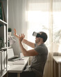 Young man using augmented VR headset at home office during a lockdown because of Covid-19. Side view Male in VR headset. Domestic life, natural backlight from window