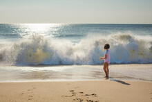 A Small Girl Stands At Edge Of Ocean Watching Wave Break Onto Beach
